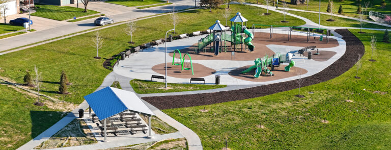 Aerial view of a modern playground with green and blue equipment, surrounding benches, and a sheltered picnic area on a circular rubber surface.