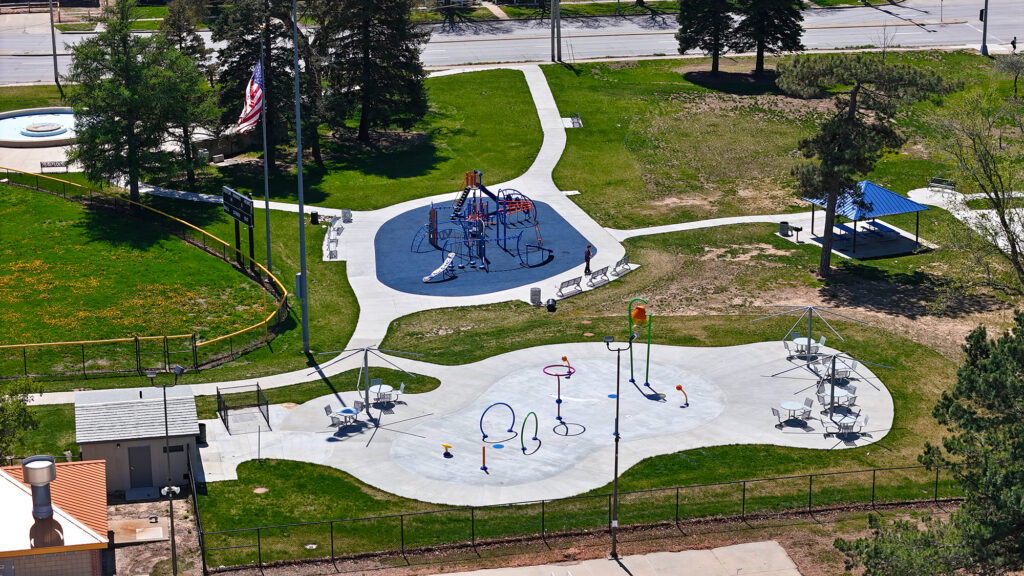 Aerial view of a park with a blue playground surface, climbing structures, and a circular splash pad surrounded by walkways and benches.