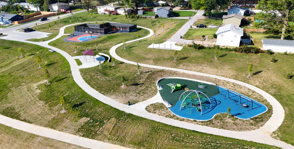 Aerial view of a community park showing playground with blue safety surface, a fenced court, curved walking paths, and surrounding houses and lawns.