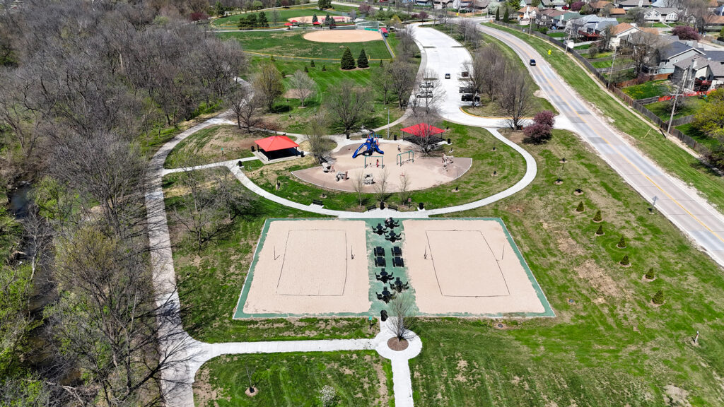 Aerial view of a park with a playground, red-roofed pavilion, circular walking paths, and surrounding trees, road, and houses nearby.