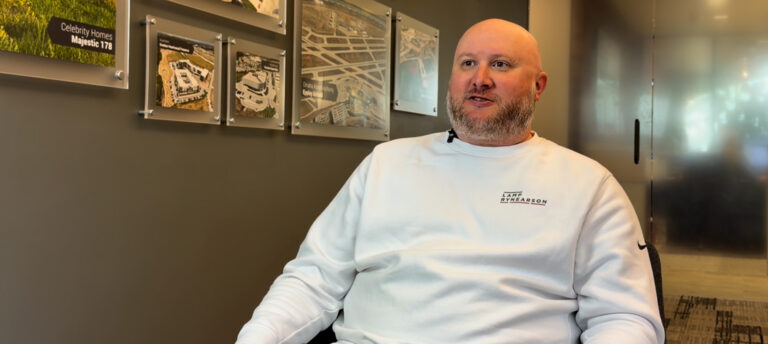 Bald man with a beard wearing a white sweatshirt sits in a modern office, with framed maps on a gray wall behind him.